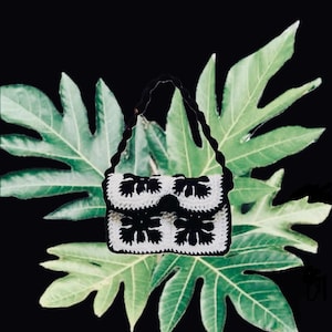 May include: A handmade crochet bag with a black and white floral design. The bag features a flap closure and a braided handle. The background is a black surface with green leaves.