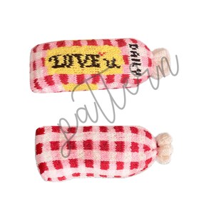 May include: Two knitted hair accessories shaped like bottles. One has a yellow label with "LOVE U" and "DAILY" text. The other is red and white checkered. Both have a cream-colored, ruffled end.