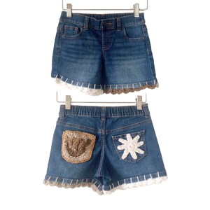 May include: Blue denim shorts with white and brown crochet trim. The shorts have a crochet patch on the back pocket. The shorts are hanging on a white hanger.