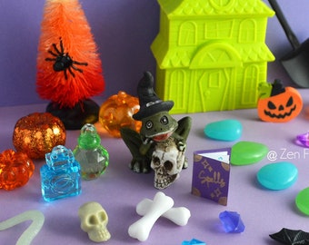 May include: A collection of Halloween-themed miniature figurines and decorations, including a green house, a frog holding a skull, a spider web with spiders, a pumpkin, a witch's hat, a book, a snake, bones, and various colored gems.