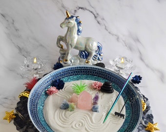 May include: A blue and white mosaic zen garden with white sand, a rake, stones, and crystals. A pink crystal is in the center of the garden. The garden is surrounded by a white lace doily and a string of lights with stars and crescent moons. A white unicorn figurine is in the background.