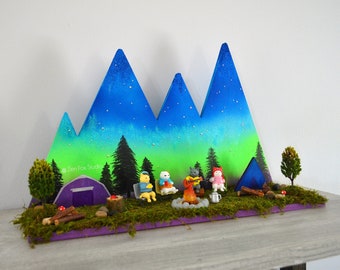May include: A miniature woodland scene with a purple tent, a campfire, and four animal figurines in front of a blue and green mountain backdrop with stars. The scene is set on a purple base with moss and other natural elements.