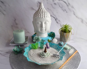 May include: A white ceramic Buddha head figurine sits on a blue and white base with a small green plant and a white sand zen garden in front of it. The zen garden includes a rake, a small wooden stick, and a few crystals.
