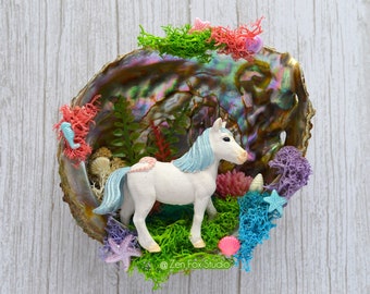 May include: A white unicorn figurine with a blue mane and tail is placed inside a large iridescent seashell. The seashell is decorated with green and pink moss, blue and purple coral, and starfish. The text "@Zen Fox Studio" is visible at the bottom of the image.
