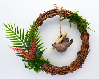 May include: A decorative wreath made of woven brown twigs, adorned with green and red faux foliage. A small, brown sloth figurine hangs from a wooden branch in the center. The wreath is set against a white background.