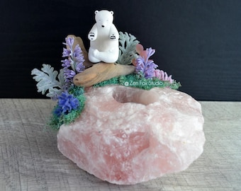 May include: A pink rose quartz candle holder with a white bear figurine in a meditative pose. The candle holder is decorated with lavender, blue, and green floral accents. The background is dark.