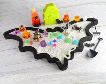 May include: A black bat-shaped tray with white sand, a green house, a witch, a spider, a skull, a rake, a shovel, and other Halloween-themed decorations.
