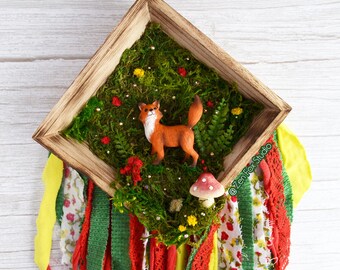 May include: A wooden framed wall hanging featuring a miniature orange fox figurine in a mossy woodland scene. The scene includes a mushroom, red and yellow flowers, and decorative fabric streamers in red, green, and floral patterns.