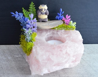 May include: A pink rose quartz candle holder with a circular opening for a tea light. The holder is decorated with blue and purple flowers, green moss, and a small raccoon figurine. The text "@Zen Fox Studio" is visible.