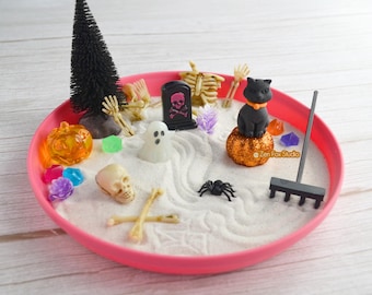 May include: A miniature Halloween-themed zen garden in a pink tray. The garden features a black cat on a glittery pumpkin, a tombstone, a skeleton, a ghost, a spider, and a small rake. The sand is white, and there are colorful gems.
