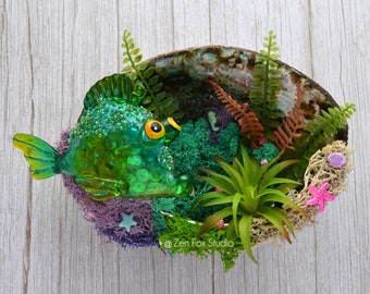 May include: A green glass fish with glitter and yellow eyes is nestled in a seashell with moss, ferns, and other sea-themed decorations. The seashell is on a light wood background.  @Zen Fox Studio