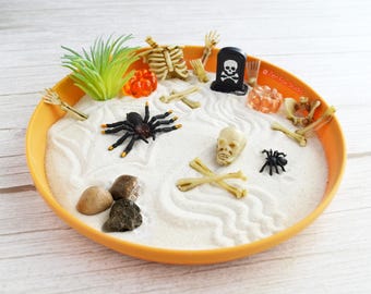 May include: An orange dish filled with white sand, decorated with Halloween-themed miniatures. The scene includes a black spider, a skull, bones, a tombstone, and small rocks. A green plant and orange gems add color to the spooky display.
