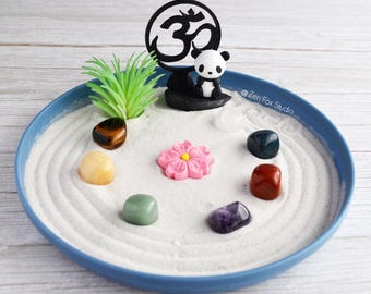 May include: A miniature zen garden in a blue dish. The garden features white sand, a black Om symbol, a panda figurine, a pink flower, green plants, and colorful stones. The text "@Zen Fox Studio" is visible.