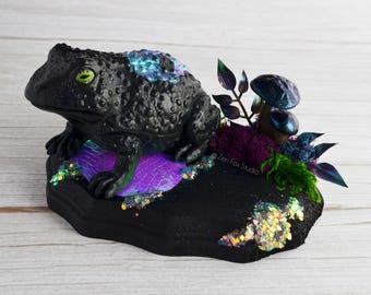 May include: A black frog figurine with iridescent blue and green accents sits on a black base with purple and iridescent glitter. The base also features a small green plant and a blue and purple mushroom.