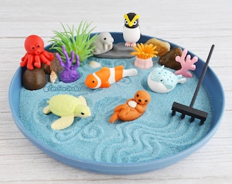 May include: A miniature zen garden with a blue sand base and various sea creature figurines. The scene includes an octopus, clownfish, turtle, penguin, and other marine life, along with a small rake and decorative plants.