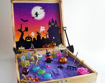 May include: A wooden box with a purple sand zen garden inside. The box has a Halloween theme with a black cat, a witch, a haunted house, and a full moon. The box also includes a rake, a shovel, and other Halloween-themed decorations.