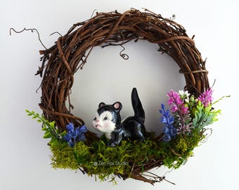 May include: A decorative wreath made of intertwined brown twigs, featuring a ceramic skunk figurine. The skunk is black and white, nestled among green moss, purple, and blue flowers. The wreath is a charming home decor item.