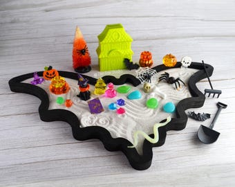 May include: A black bat-shaped sand tray with white sand and Halloween-themed decorations, including a witch, a house, pumpkins, a spider, a skull, and a snake. There are also colorful gems and stones in the tray.
