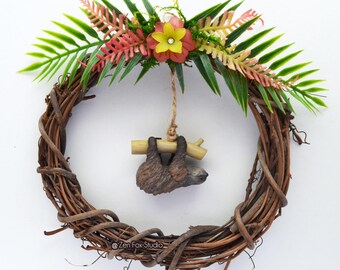 May include: A handcrafted grapevine wreath adorned with faux greenery, pink and yellow flowers, and a small sloth figurine hanging from a branch. The wreath is a circular shape and is suitable for indoor or outdoor use.