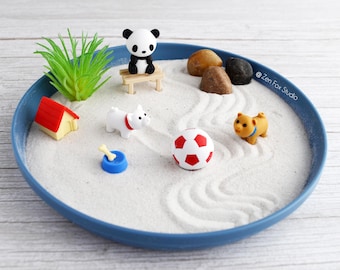 May include: A miniature zen garden with a blue tray filled with white sand. It features a panda on a bench, a soccer ball, a dog, a house, rocks, and a small plant. The sand is raked with wave patterns.