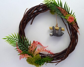 May include: A handcrafted wreath made of brown twigs, adorned with green and red faux foliage, and a small gray sloth hanging from a wooden bar. The wreath is decorated with a small orange flower and green moss.