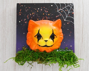 May include: A Halloween-themed orange and yellow cat face with black eyes and a black nose, surrounded by green moss and a black background with white glitter and a white spider web.