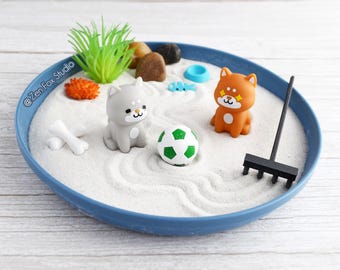 May include: A miniature zen garden with a blue rim. The sand-filled garden features two Shiba Inu figurines, a soccer ball, a rake, and other decorative elements. The garden includes the text "@ZenFox Studio".