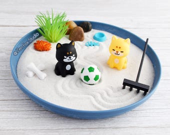 May include: A miniature zen garden in a blue tray features a black and yellow Shiba Inu figurine, a soccer ball, and a rake. The garden includes white sand with raked patterns, small rocks, and a bone. The text "@Zen Fox Studio" is visible.