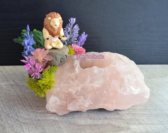 May include: A pink rose quartz crystal candle holder with a small lion figurine in a meditative pose, surrounded by colorful artificial flowers and greenery. The candle holder is on a light gray surface.