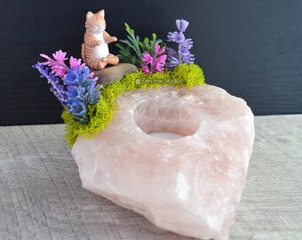 May include: A pink rose quartz candle holder with a carved-out center for a tea light. The holder is decorated with a small figurine of a cat, faux flowers, and green moss. The cat is sitting on a small rock. The background is black and gray.