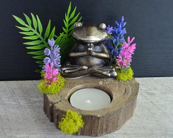 May include: A bronze-colored frog figurine in a meditative pose sits atop a wooden candle holder. The holder features a tealight candle, green fern, and colorful faux flowers. The design includes a rustic, natural aesthetic.
