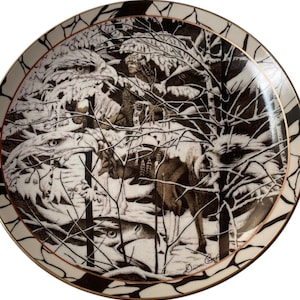 May include: A decorative plate with a detailed black and white illustration of a Native American figure, eagles, and deer in a snowy forest scene. The plate has a gold rim and a patterned border.