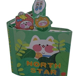 May include: A green pop-up card with a cartoon cat and the words "NORTH STAR". The card features illustrations of cartoon characters in costumes, including a strawberry, a flower, and a pair of birds on skewers.