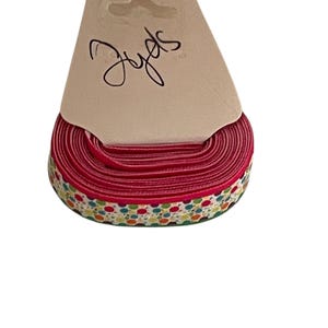 May include: A spool of red ribbon with a patterned edge featuring small, colorful dots. The ribbon is attached to a tan card with the handwritten word "Jyds" in black script.