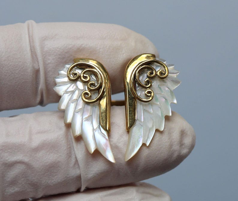 May include: A pair of angel wing earrings. Each earring features a gold-colored metal frame with a swirling design and iridescent, white, feather-like details. The earrings are designed to resemble a pair of wings.