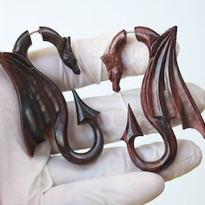 May include: A pair of wooden dragon ear gauges, one with a dark brown finish and the other with a reddish brown finish. The dragon wings are carved with intricate details and the tails are curled into a hook shape.