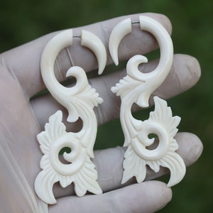 May include: A pair of white bone earrings with a carved floral design. The earrings are shaped like a curved stem with a flower at the bottom.