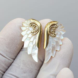 May include: A pair of gold-colored wing-shaped earrings with iridescent mother-of-pearl feathers. The earrings have a decorative gold filigree design at the top. The earrings are designed to be worn as a pair.