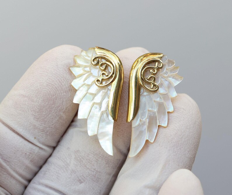 May include: A pair of gold-colored wing-shaped earrings with iridescent mother-of-pearl feathers. The earrings have a decorative gold filigree design at the top. The earrings are designed to be worn as a pair.