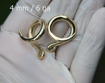 May include: Two gold-colored, spiral-shaped ear gauges. The gauges are 4 mm / 6 ga. The gauges are held in a gloved hand. The gauges have a smooth, polished finish and a curved design.