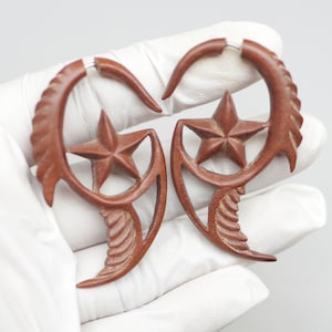 Op de afbeelding: Een paar houten oorbellen met een stervormig ontwerp. De oorbellen zijn bruin en hebben een natuurlijke houten afwerking. Ze zijn gevormd als een halve maan met een ster in het midden.