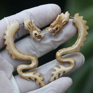Op de afbeelding: Een paar drakenoorbellen van been. De oorbellen zijn lichtbruin van kleur en hebben ingewikkelde details. Ze worden getoond op een wit oppervlak.