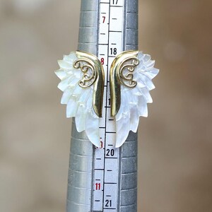 May include: A unique ring featuring a carved white mother-of-pearl design with gold-colored accents. The ring's design resembles a stylized leaf or wing shape. The ring is being measured on a ring sizer.