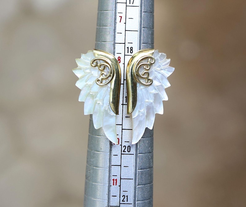 May include: A unique ring featuring a carved white mother-of-pearl design with gold-colored accents. The ring's design resembles a stylized leaf or wing shape. The ring is being measured on a ring sizer.