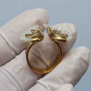 May include: A gold-toned ring with a unique design. The ring features two white, fan-shaped elements with gold filigree accents. The ring is held by a gloved hand against a light blue background.