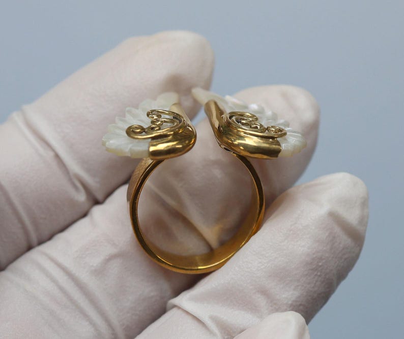 May include: A gold-toned ring with a unique design. The ring features two white, fan-shaped elements with gold filigree accents. The ring is held by a gloved hand against a light blue background.