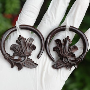 Op de afbeelding: Een paar donkerbruine houten oorbellen. Elke oorbel heeft een gesneden bloemenontwerp aan de basis van een cirkelvormige hoepel. De oorbellen worden in een gehandschoende hand gehouden tegen een vage groene achtergrond.