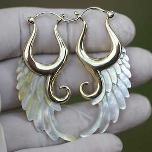 Puede incluir: Un par de pendientes de aro dorados con diseño de alas. Las alas están hechas de material iridiscente, en capas, blanco y crema. Los pendientes tienen una forma curva y elegante.
