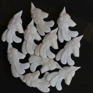 May include: A collection of off-white carved wolf head pendants. Each pendant features detailed feather accents and a circular design near the ear. The pendants are arranged on a black surface, showcasing their intricate craftsmanship.