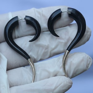 Op de afbeelding: Een paar zwarte, gebogen oorbellen met zilverkleurige punten. De oorbellen hebben een haakontwerp en zijn gemaakt van een donker materiaal, mogelijk hout. Ze worden getoond tegen een lichte achtergrond.